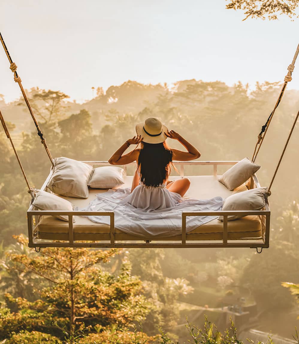 Female tourist on holiday admiring the view while sitting on outdoor hanging bed elevated above the tropical forest in Bali, Indonesia